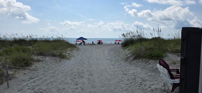 The path through the dunes opens up to umbrellas and beach chairs waiting on the sand.