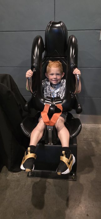 One of the kids beams from inside a padded ride seat, harness buckled and grips in hand, ready for launch.
