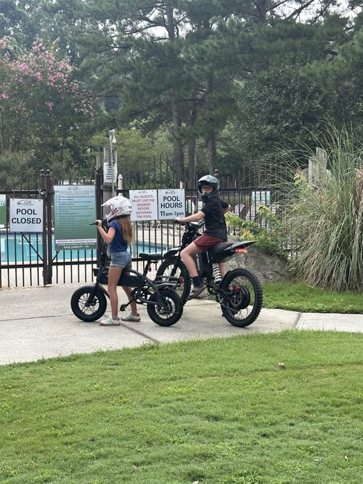Helmets on, the two kids straddle their e-bikes outside the still-closed pool gate.