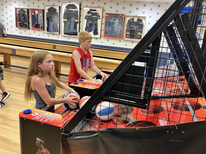 The kids team up on the arcade basketball machine, framed by a wall of vintage badges and dress uniforms.