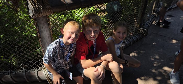 The kids pile together on the rope fence, sunlight streaking through the jungle canopy behind them.