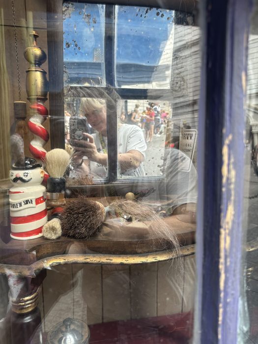 A vintage barbershop display of shaving brushes and mugs glimpsed through an old shop window.