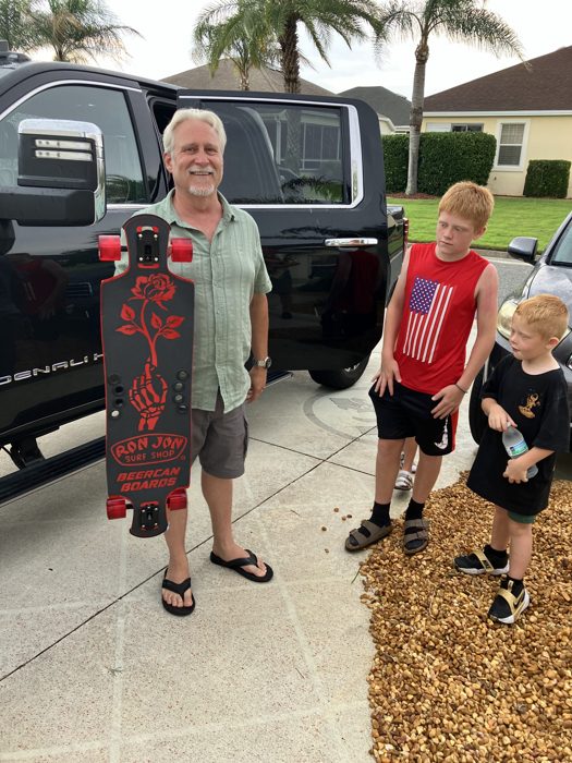 Another angle on the new longboard show-and-tell, the kids clustered close in the driveway.