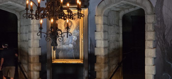 Candlelit chandeliers glowing in front of the mad scientist's portrait at the queue entrance.