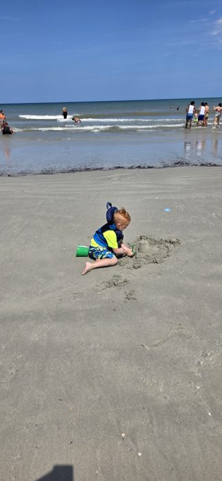 A small sandcastle takes shape in the foreground while other beachgoers wade in the surf beyond.