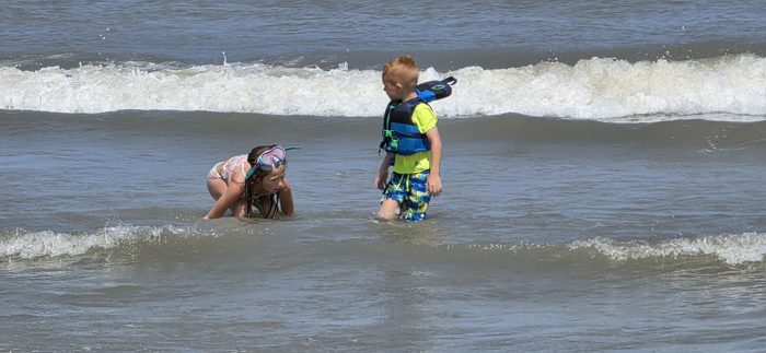 One of the kids crouches low to check out something in the shallows while a sibling in a life vest looks on.