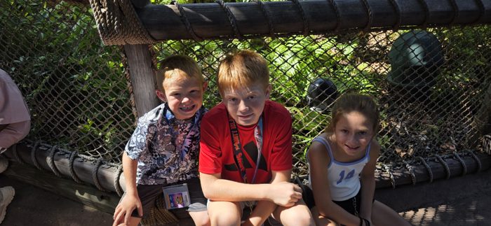 The kids huddle together on the rope-lined bench in the shady queue, all grins for the camera.