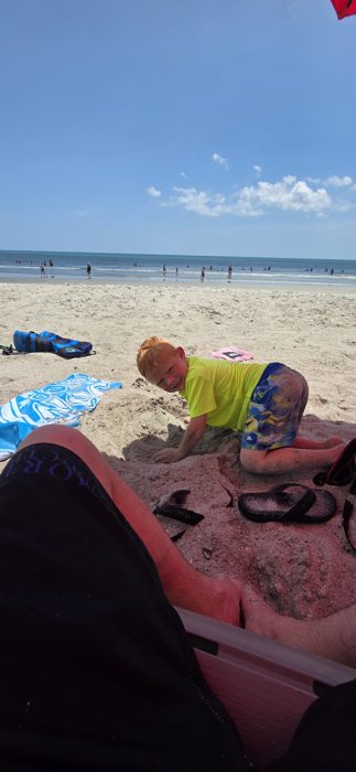 One of the kids grinning up from a sandy dig, sunglasses and flip-flops scattered nearby.