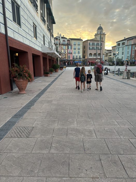 The family strolls the waterfront path toward the resort harbor as the sun sets behind the pastel towers.