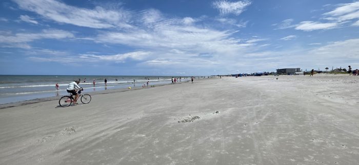 A cyclist pedals along the hard-packed sand while swimmers cool off in the surf.
