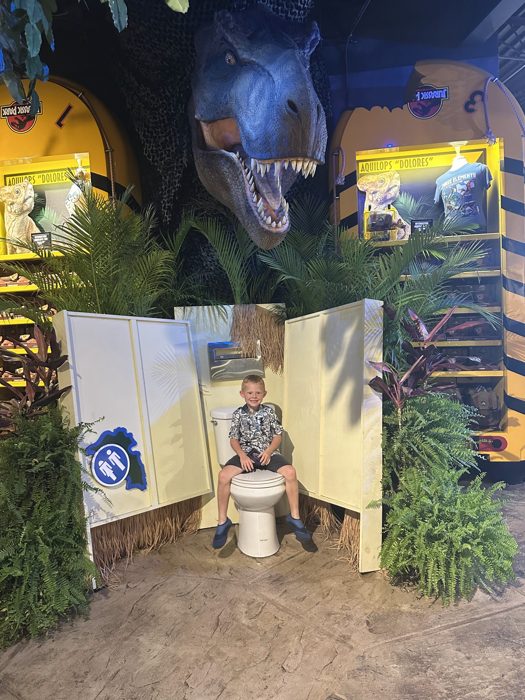 One of the kids grins from the jungle outhouse photo op, a towering T-rex head looming in from above.