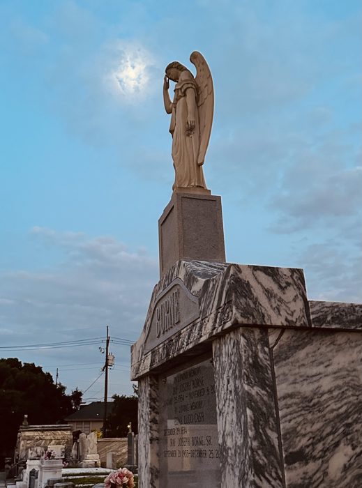 A weeping angel statue silhouetted against a sunburst sky atop a tomb.