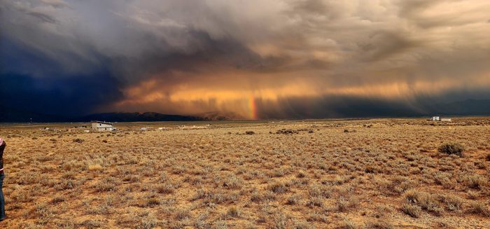 A wall of monsoon rain sweeps across the high desert in golden light, with our little camper dwarfed beneath the storm.