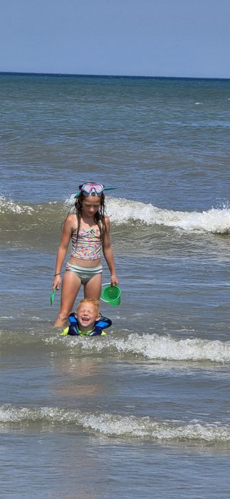 One of the kids beams through the ocean spray as a sibling wades past with snorkel gear and a bright green sand bucket.