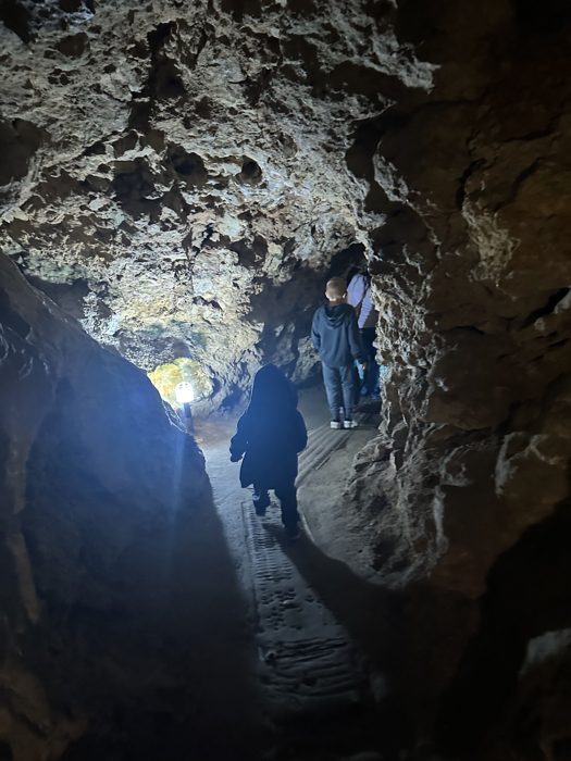 A lone figure descends a narrow lit cave corridor far underground.