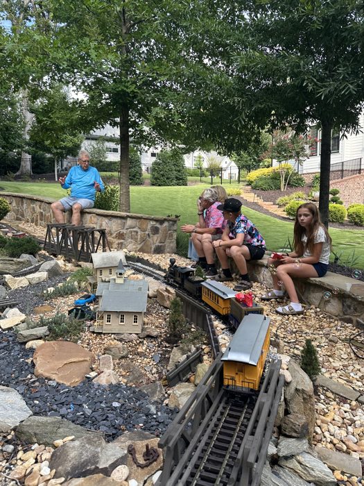 The family gathers along the garden wall to watch the trains roll through, bubbles drifting over the miniature town.