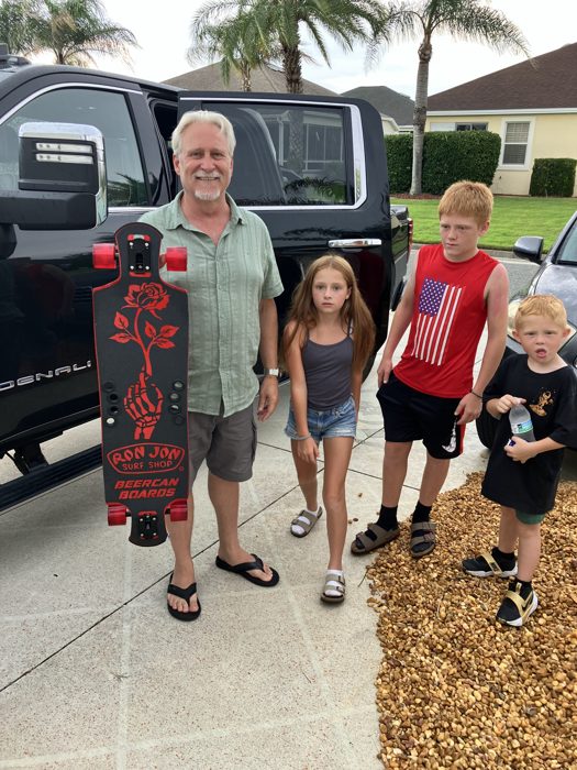 A family member proudly holds up a new Ron Jon longboard while the kids gather around in the driveway.