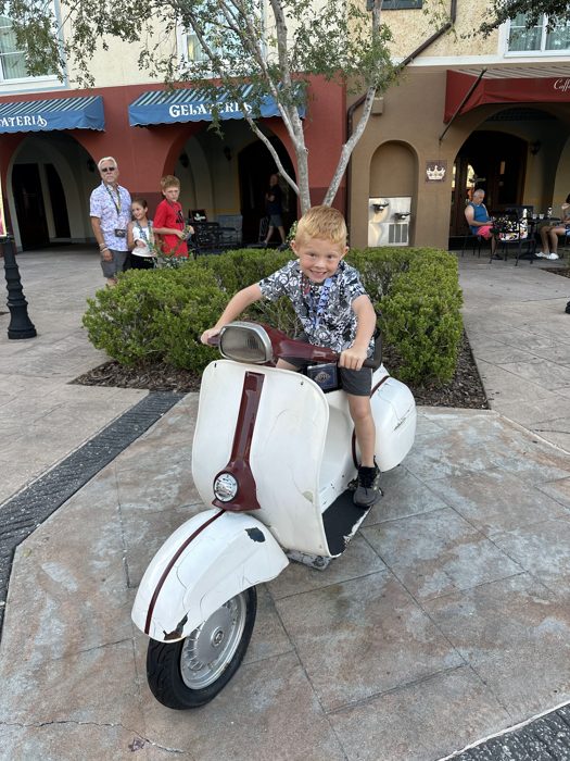 One of the kids gives a thumbs-up while straddling a vintage scooter statue in the Italian-style courtyard, the rest of the family looking on.