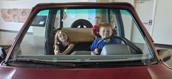 Kids grinning from behind the wheel of the vintage car exhibit.