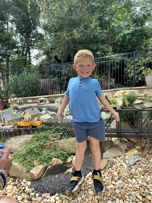 One of the kids beams next to the miniature train village winding through the rock garden.