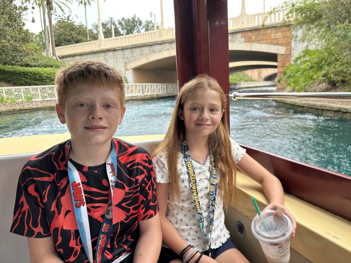 The kids ride the water taxi together, lanyards swinging, as a stone bridge slides past behind them.