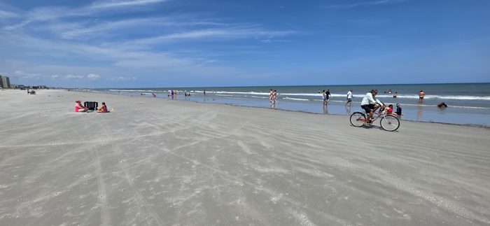 Another cyclist rolls past as beachgoers spread out along the shoreline in the afternoon sun.