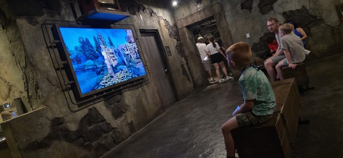 One of the kids waits it out on a bench inside the ride queue, a spooky garden scene glowing on the screen ahead.