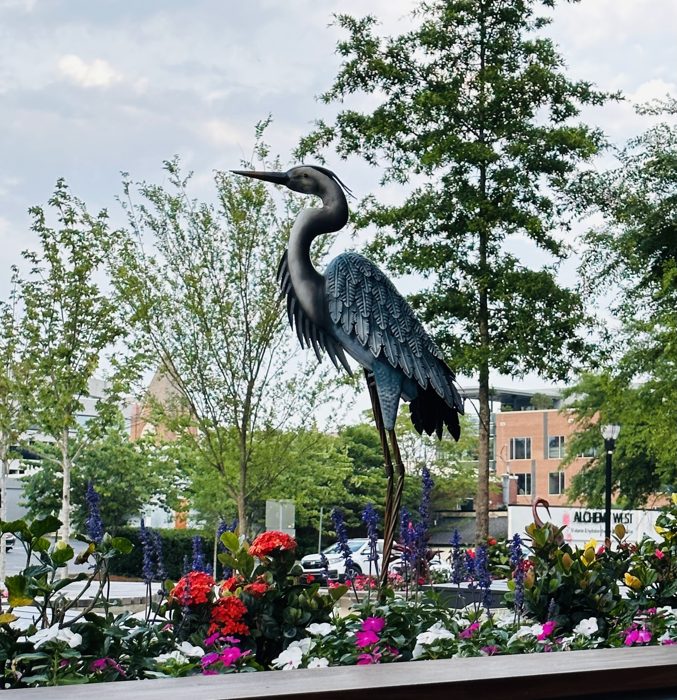 A tall blue heron sculpture standing amid the flowering gardens.