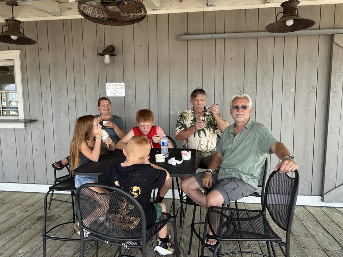 The family digs into ice cream cups together on a shaded porch.