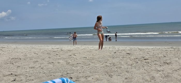 One of the kids stands watching the waves while others wade and carry a bodyboard back up the beach in the distance.