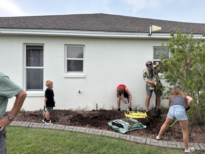 The whole crew pitches in on a flower bed refresh, shovels and bags of garden soil scattered across the yard.