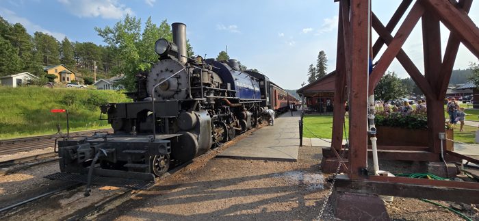 The big black steam locomotive rests beneath the timber trestle as the crowd gathers at the depot.