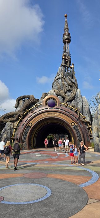 Sunlight breaks over the mosaic plaza as guests stream beneath the towering stone archway into the monster-themed land.