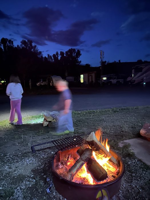 Blue-hour over the campground firepit as the kids circle a roaring evening fire.