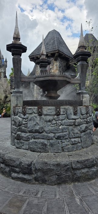 A smoking cauldron fountain rises from old stone in the Wizarding World's village square.