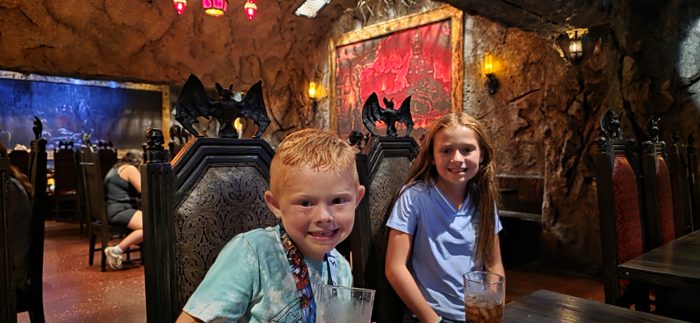 The kids grin over their drinks at a candlelit table, carved bats watching from the booth behind them.