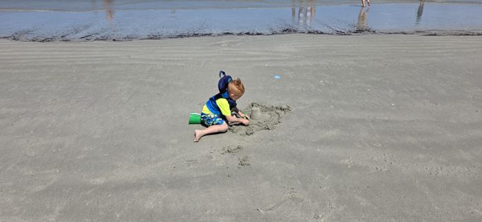 Focused sandcastle building on the shore as beachgoers pass by in the background.