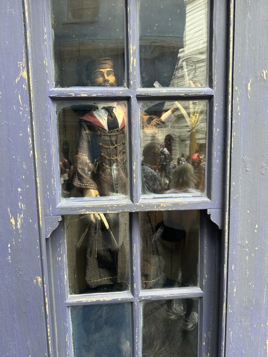 A wizard mannequin peers out from behind a shop's dusty, cracked-paint window display.